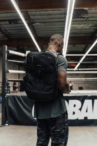 Man wearing the Midnight Black Ai1 backpack on both shoulders in a boxing gym, rear view