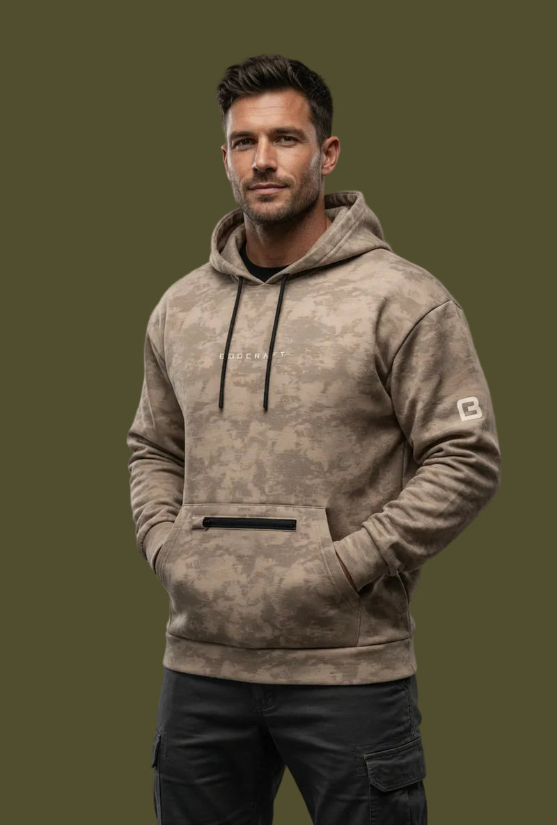 Man wearing a brown camouflage hoodie with a brand logo, standing against a concrete wall.