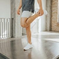 Pebble Grey men's golf shorts mid quad stretch, heel pulled up toward the hip in a loft studio
