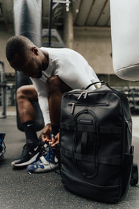 Athlete lacing up shoes next to the Midnight Black Ai1 gym backpack in a boxing gym