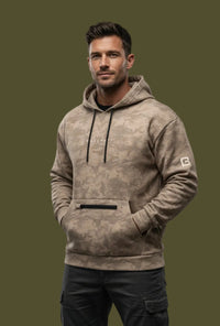 Man wearing a brown camouflage hoodie with a brand logo, standing against a concrete wall.